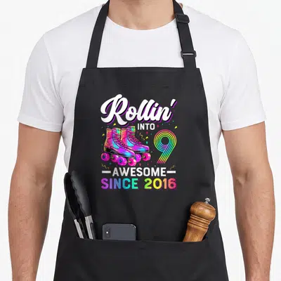 Nine roller birthday apron - Rollin into 9 awesome since 2016 rainbow roller skates cartoon graphic — shown on a black cotton canvas apron against a white background
