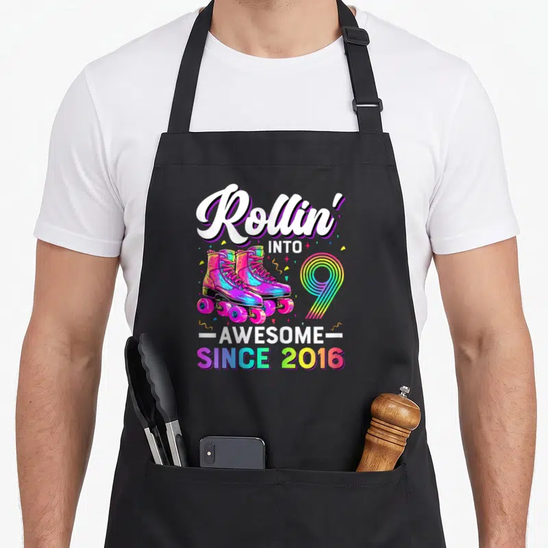 Nine roller birthday apron - Rollin into 9 awesome since 2016 rainbow roller skates cartoon graphic — shown on a black cotton canvas apron against a white background