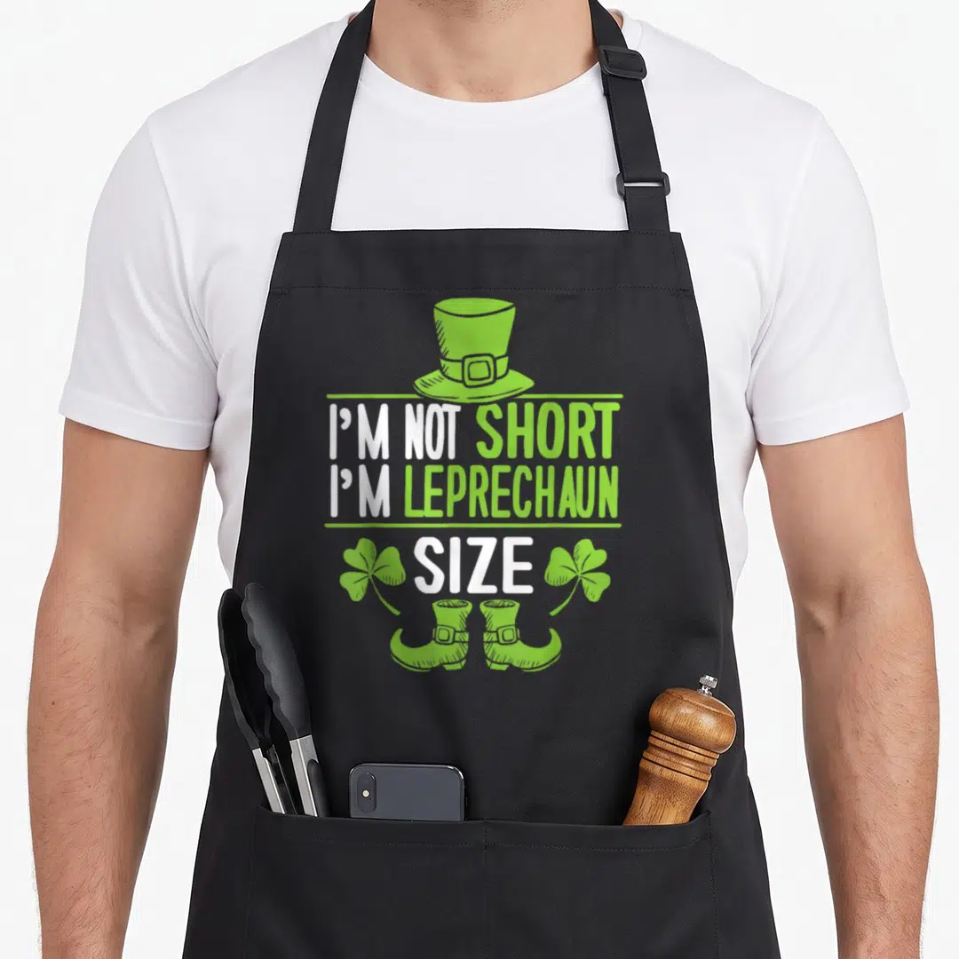 Retro leprechaun st patrick day aprons - Green and white cartoon leprechaun hat, boots, shamrocks, and stacked text — shown on a black cotton canvas apron against a white background
