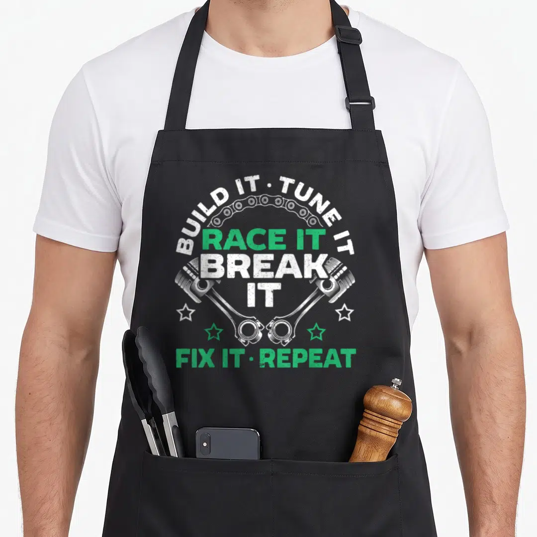 Build adrenaline apron - Crossed pistons and chain arch with stacked racing text in white and green distressed style — shown on a black cotton canvas apron against a white background