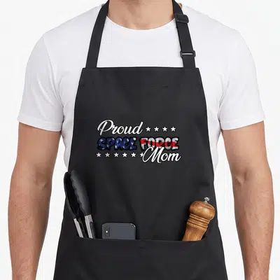 Bold force mom apron - Stacked Proud Space Force Mom text with red white and blue flag fills and nine stars — shown on a black cotton canvas apron against a white background