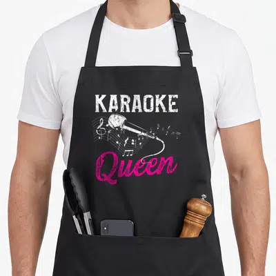 Night queen apron - Distressed white karaoke text, pink Queen script, and microphone with musical notes on black — shown on a black cotton canvas apron against a white background