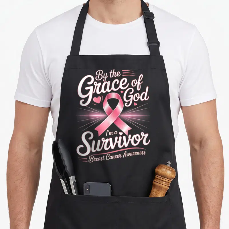 Survivor grace apron - Stacked white lettering and ribbon graphic with small hearts — shown on a black cotton canvas apron against a white background