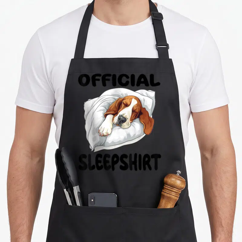 Hound sleepshirt dog apron - Cartoon basset hound sleeping on pillow between official sleepshirt text — shown on a black cotton canvas apron against a white background