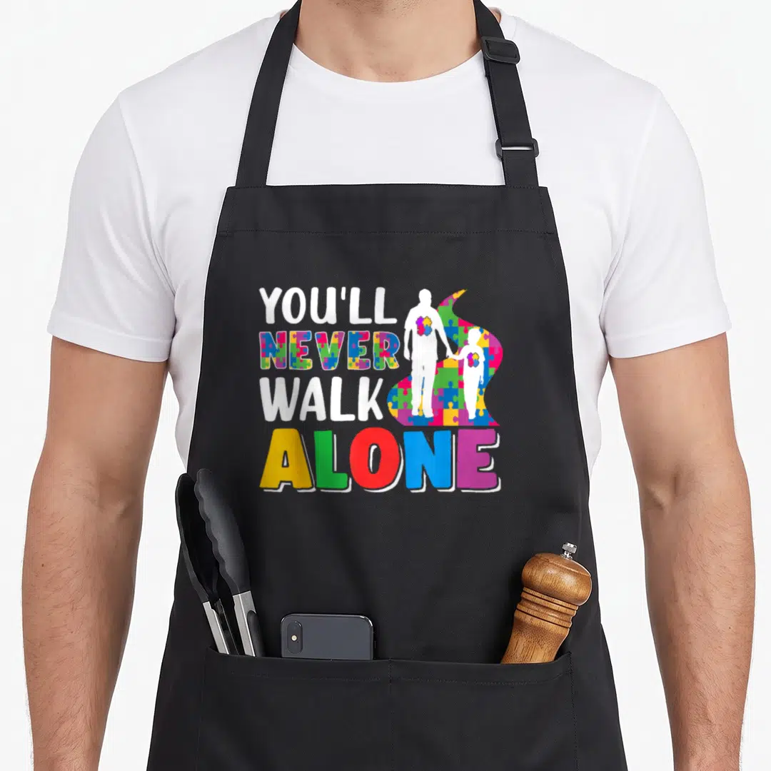 Youll father son apron - YOU'LL NEVER WALK ALONE text with colorful puzzle silhouettes of a man and child holding hands — shown on a black cotton canvas apron against a white background