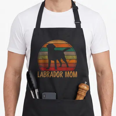 Mother labrador apron - White dog silhouette over striped retro sun with orange LABRADOR MOM text below — shown on a black cotton canvas apron against a white background