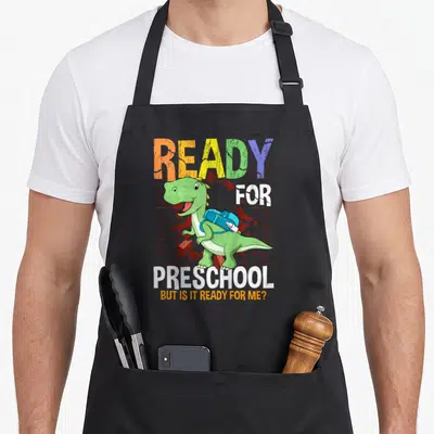 Toddler preschool apron - Green cartoon dinosaur with blue backpack breaking red brick wall with multi-colored text — shown on a black cotton canvas apron against a white background