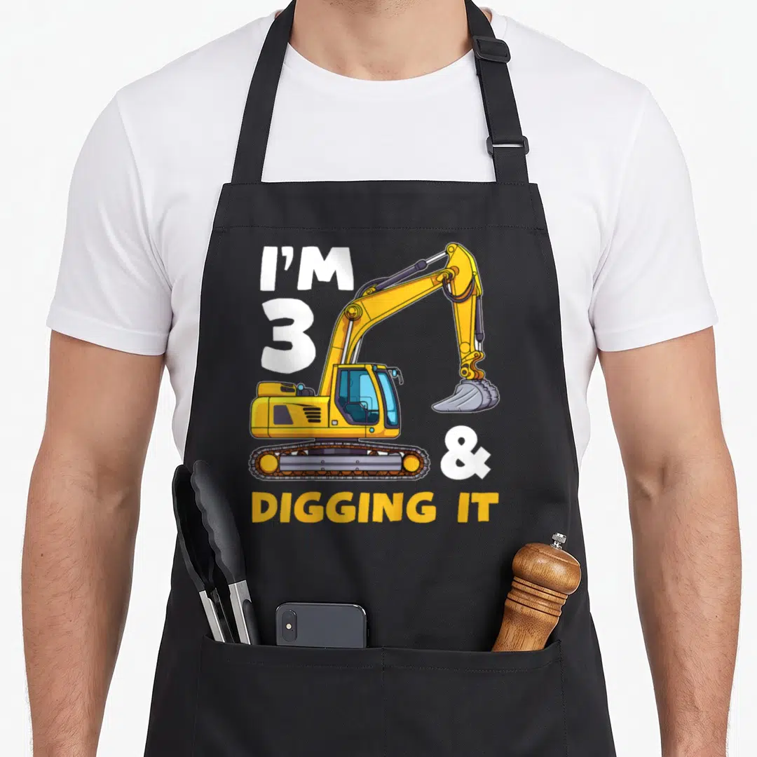 Birthday excavator truck apron - I'M 3 & DIGGING IT text beside yellow cartoon excavator with grey bucket — shown on a black cotton canvas apron against a white background