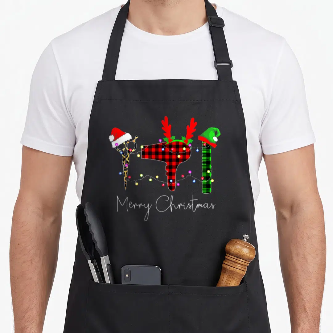 Hairstylist merry christmas apron - Salon scissors, plaid hair dryer, and green comb wearing Santa hats and antlers — shown on a black cotton canvas apron against a white background