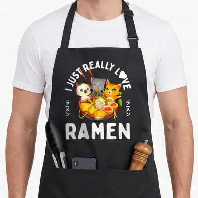 Noodle kawaii cat apron - Three cats inside a ramen bowl with I JUST REALLY LOVE RAMEN text and Japanese characters — shown on a black cotton canvas apron against a white background