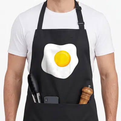 Food toast apron - Cartoon fried egg with white edges and yellow yolk centered on black background — shown on a black cotton canvas apron against a white background