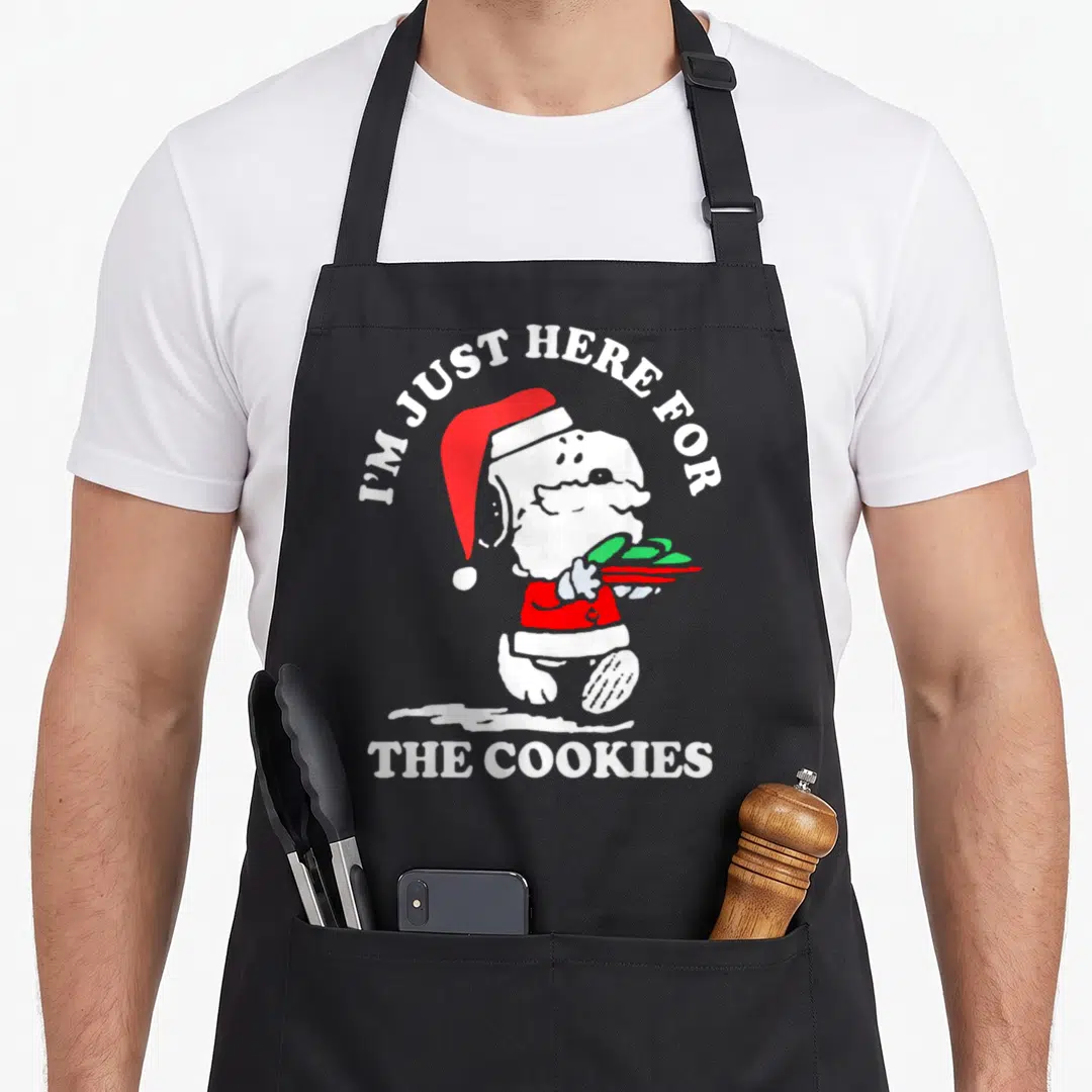 Cute cooky women's christmas aprons - Cartoon dog in red Santa suit holding green treat with white text I'M JUST HERE — shown on a black cotton canvas apron against a white background