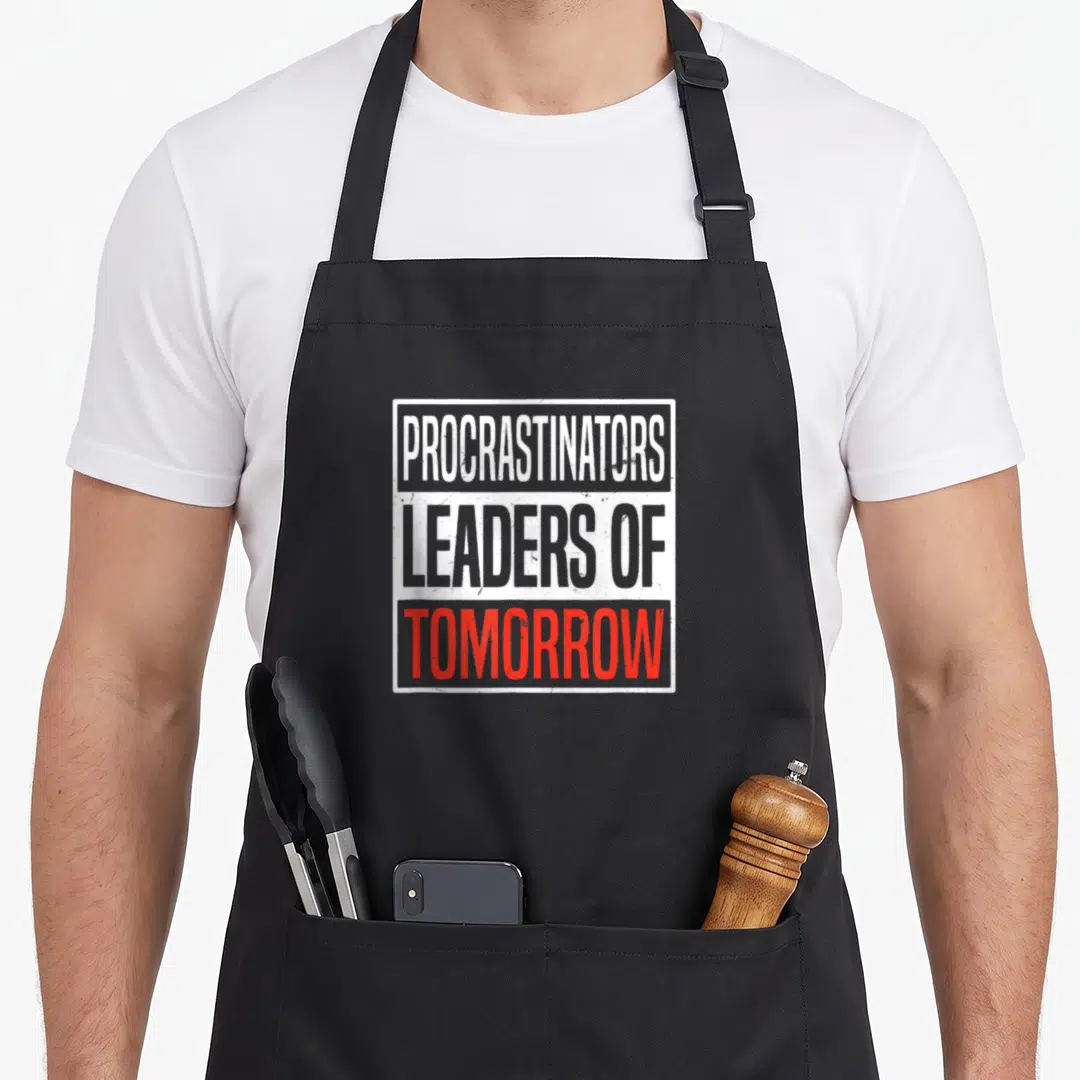 Procrastinator procrastination apron - Stacked text reading procrastinators leaders of tomorrow in white grey and red — shown on a black cotton canvas apron against a white background