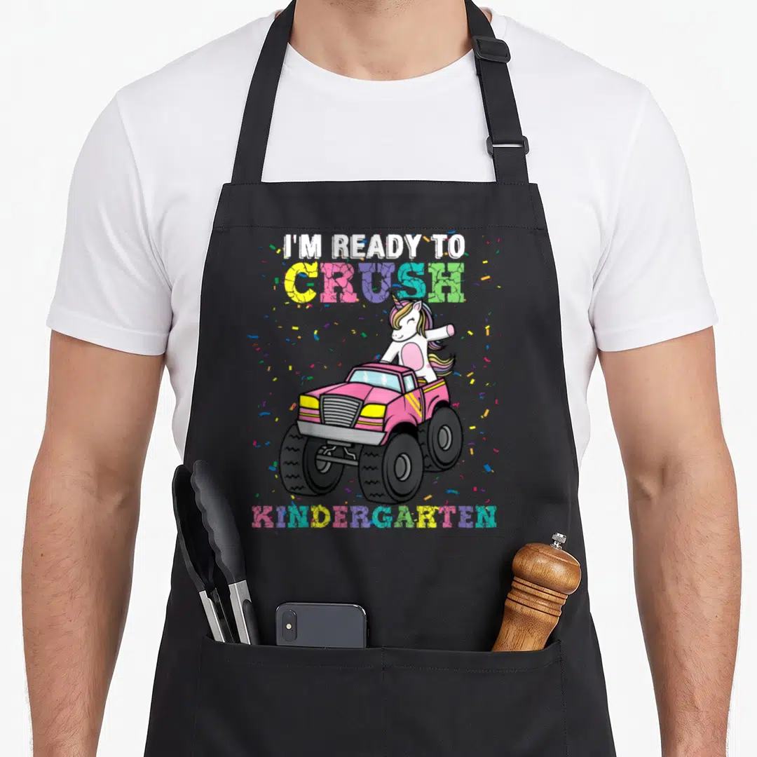 Monster unicorn aprons - Unicorn driving pink monster truck with I'm Ready to Crush Kindergarten text and confetti — shown on a black cotton canvas apron against a white background