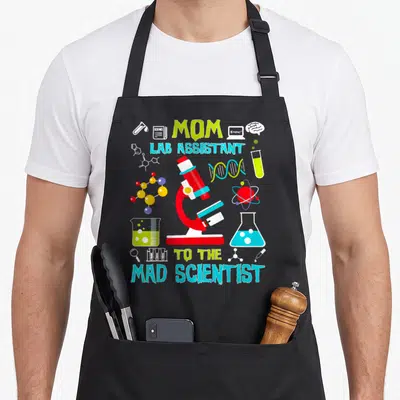 Lab birthday apron - MOM LAB ASSISTANT TO THE MAD SCIENTIST text with red microscope and colorful science icons — shown on a black cotton canvas apron against a white background