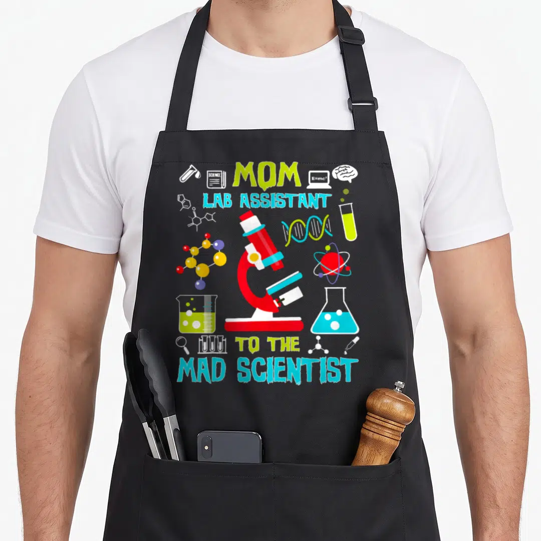 Lab birthday apron - MOM LAB ASSISTANT TO THE MAD SCIENTIST text with red microscope and colorful science icons — shown on a black cotton canvas apron against a white background