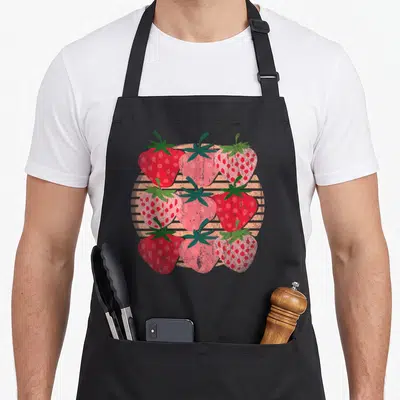 Cottagecore strawberry apron - Nine red pink and green strawberries in a grid over orange stripes — shown on a black cotton canvas apron against a white background