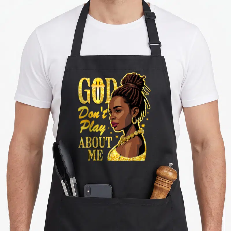 God play christian apron - Stacked gold text GOD Don't Play ABOUT ME beside cartoon portrait of Black woman with braids — shown on a black cotton canvas apron against a white background