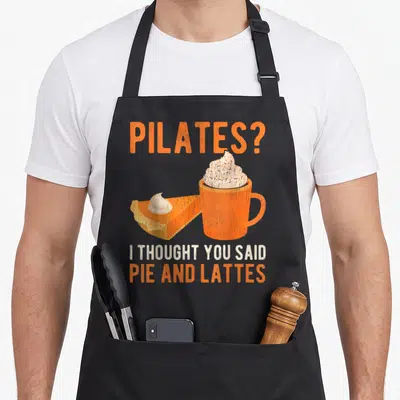Pilate coffee apron - Orange mug with whipped cream next to pie slice and Pilates pun text — shown on a black cotton canvas apron against a white background