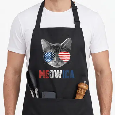 Meowica flag 4th of july apron - Grey cat wearing American flag sunglasses above distressed red white blue MEOWICA text — shown on a black cotton canvas apron against a white background