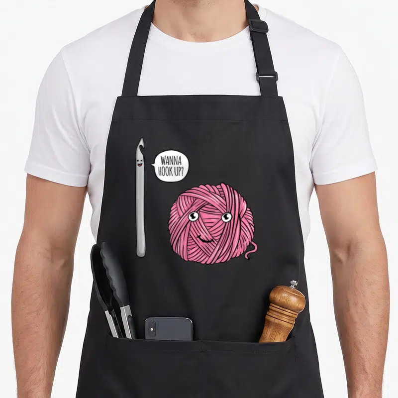 Hook crochet apron - Silver crochet hook and pink yarn ball with text Wanna Hook Up in white speech bubble — shown on a black cotton canvas apron against a white background