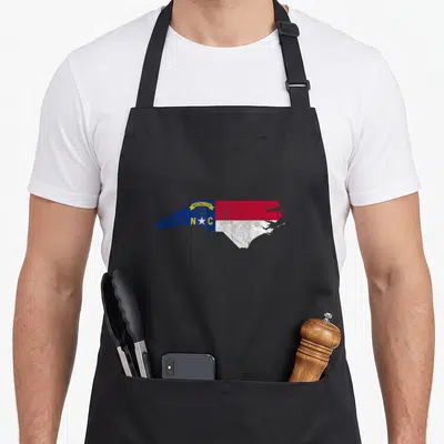 Carolina state pride apron - North Carolina map shape filled with state flag blue red white yellow and date MAY 20th — shown on a black cotton canvas apron against a white background
