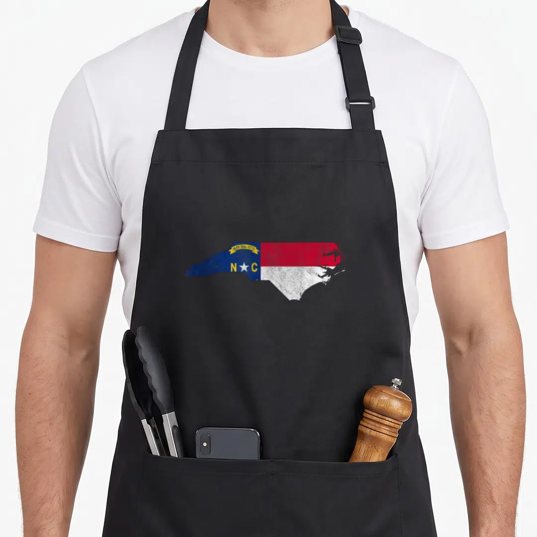Carolina state pride apron - North Carolina map shape filled with state flag blue red white yellow and date MAY 20th — shown on a black cotton canvas apron against a white background