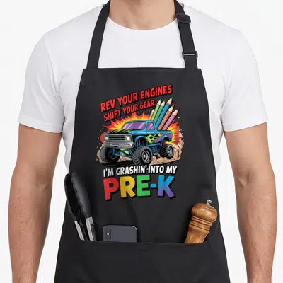 School pre truck apron - Blue monster truck with pencils and Rev Your Engines Shift Your Gear I'm Crashin' Into My — shown on a black cotton canvas apron against a white background