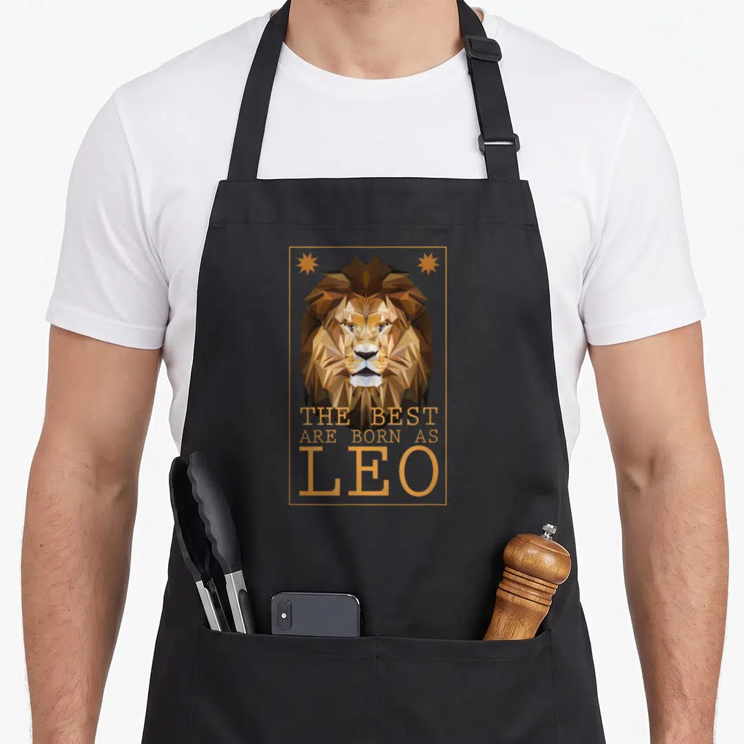 Zodiac lion apron - Geometric brown lion head in gold frame with stacked text THE BEST ARE BORN AS LEO — shown on a black cotton canvas apron against a white background