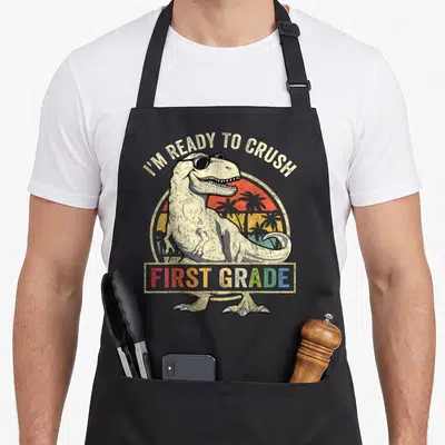 Crush dinosaur apron - T-Rex sunglasses retro sunset I'M READY TO CRUSH FIRST GRADE text arching over dinosaur — shown on a black cotton canvas apron against a white background