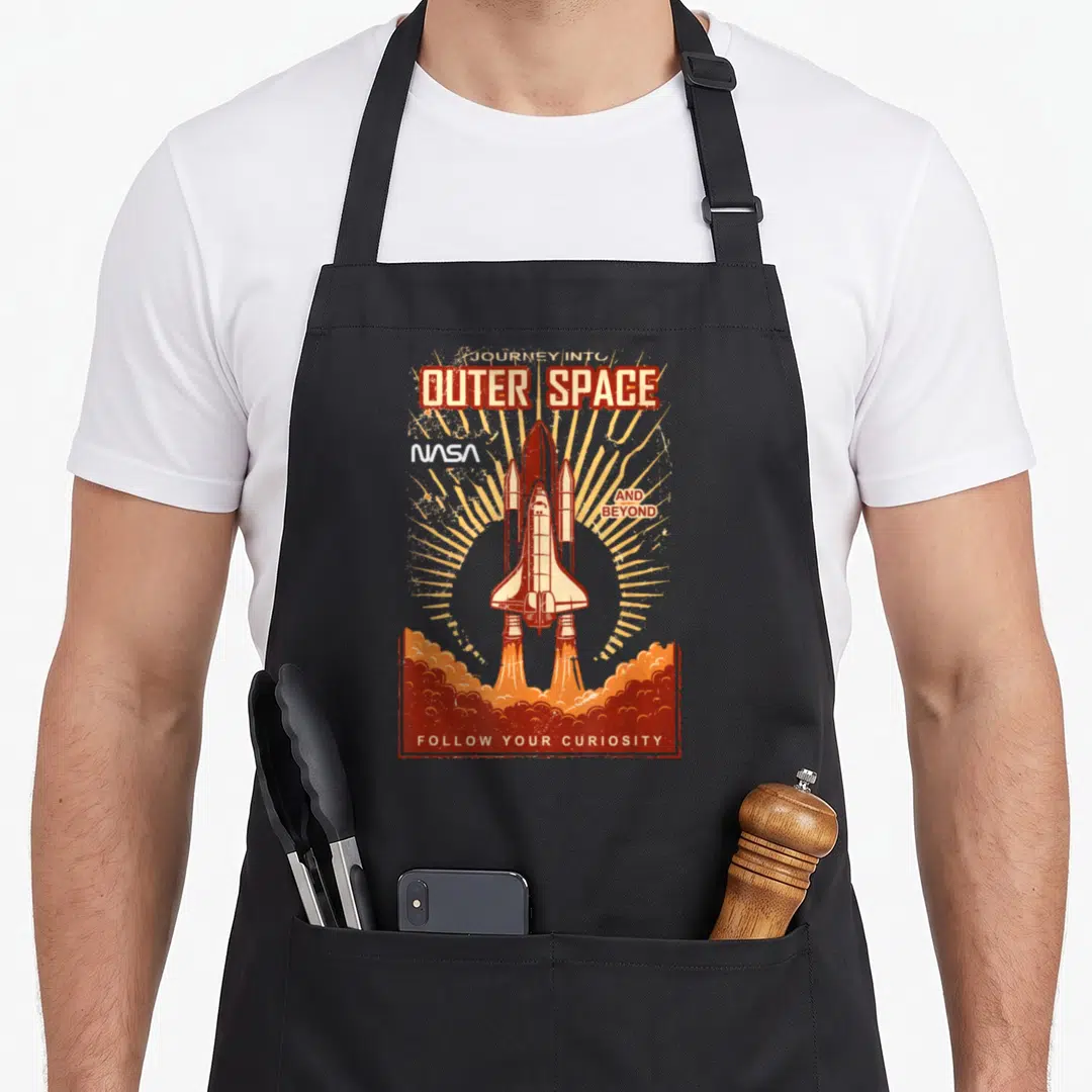 Outer space nasa apron - Retro space shuttle launch with orange smoke and text Journey Into Outer Space And Beyond — shown on a black cotton canvas apron against a white background