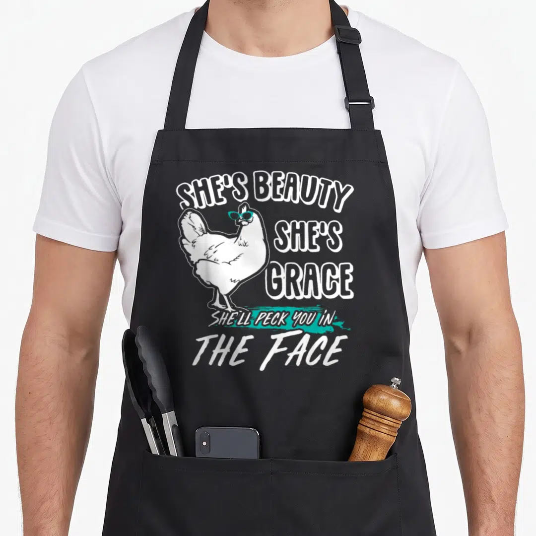 Beauty grace apron - Cartoon white chicken in blue sunglasses with text SHE'S BEAUTY SHE'S GRACE SHE'LL PECK YOU — shown on a black cotton canvas apron against a white background