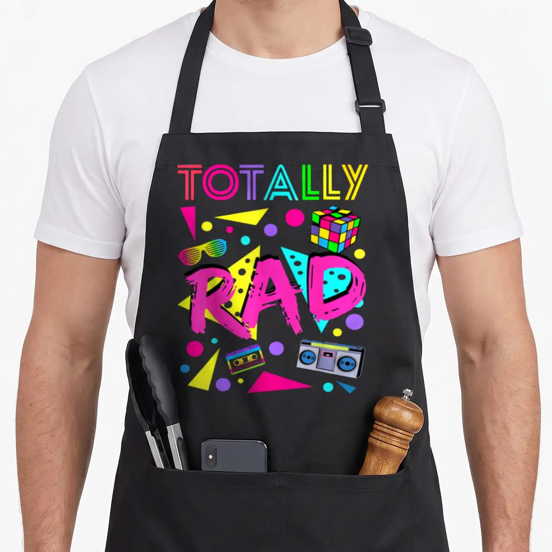 Totally 1980s apron - Totally Rad typography with boombox, cassette, shutter shades, and geometric shapes in neon colors — shown on a black cotton canvas apron against a white background