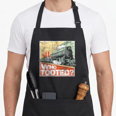 Tooted model railroad train apron - Grey steam locomotive with WHO TOOTED text on yellow and green background — shown on a black cotton canvas apron against a white background