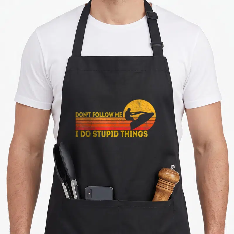Skier jetski apron - Retro jet ski silhouette over striped sun with text Don't Follow Me I Do Stupid Things — shown on a black cotton canvas apron against a white background