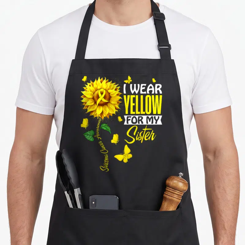 Support sarcoma awareness apron - Sunflower with ribbon and text I wear yellow for my sister sarcoma awareness — shown on a black cotton canvas apron against a white background