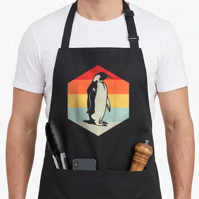 Zookeeper penguin aprons - Grey penguin standing inside a hexagon with horizontal red, orange, yellow, and light blue — shown on a black cotton canvas apron against a white background