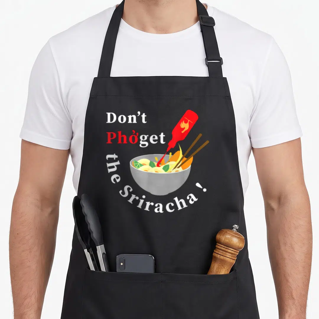 Sriracha pho apron - Cartoon pho bowl with red sauce pour and text Don't Phởget the Sriracha — shown on a black cotton canvas apron against a white background
