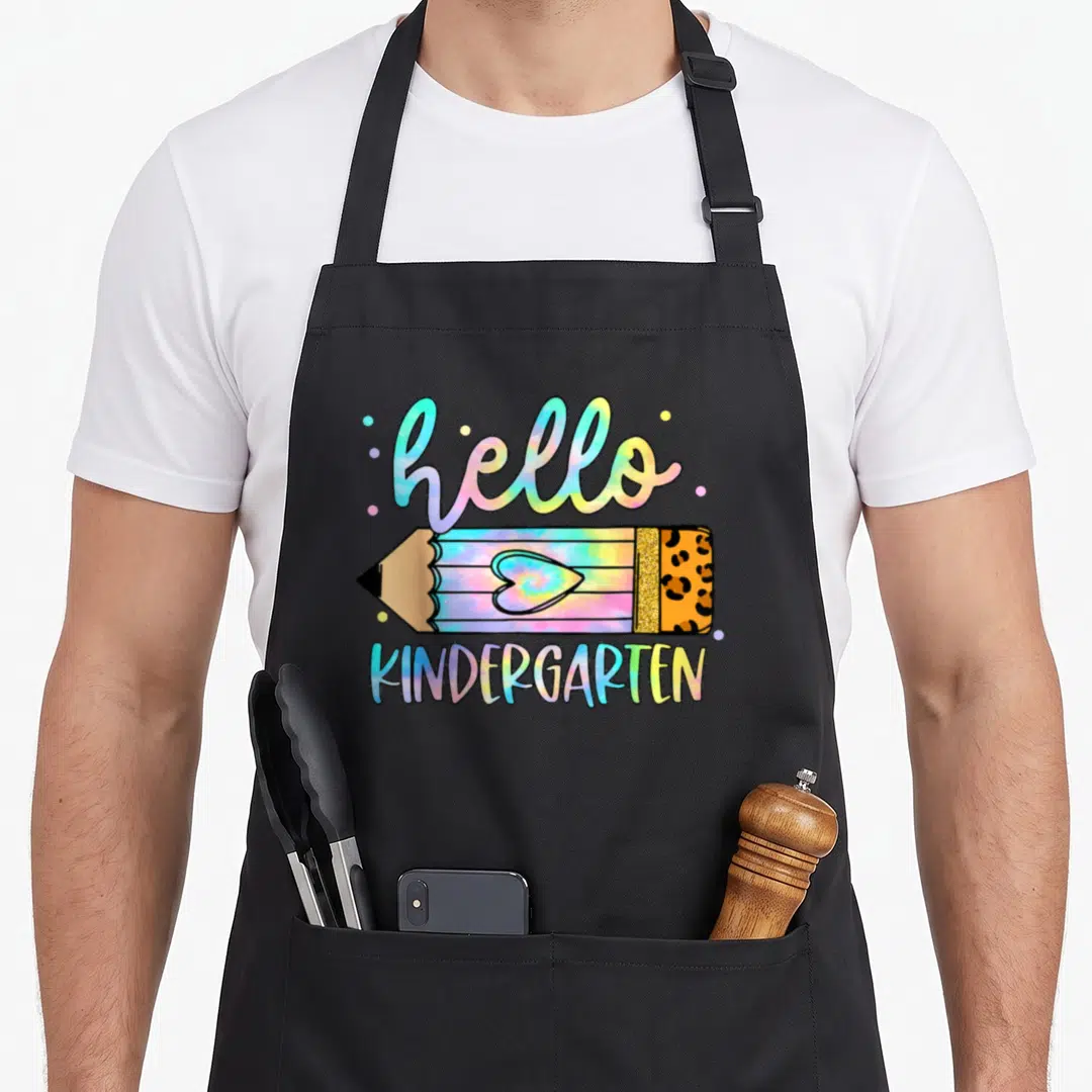 School tie-dye apron - Hello Kindergarten pencil with heart, leopard eraser, and colorful tie-dye dots — shown on a black cotton canvas apron against a white background