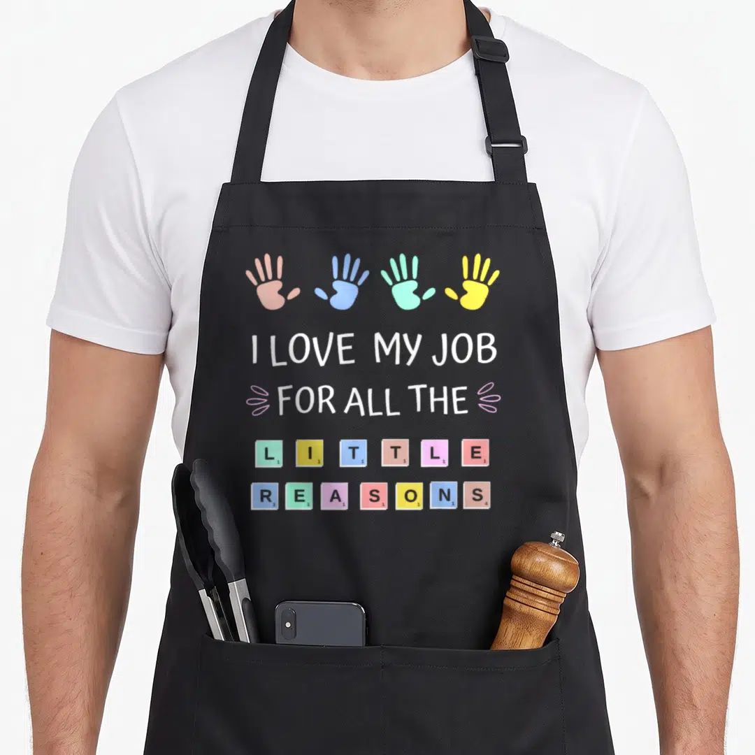 Cute reason apron - Four handprints and colorful tiles spelling I Love My Job For All The Little Reasons — shown on a black cotton canvas apron against a white background