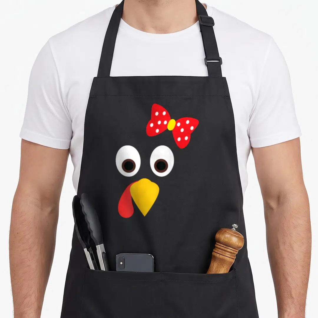 Turkey face thanksgiving apron - Cartoon turkey face with red polka dot bow, two white eyes, yellow beak, and red wattle — shown on a black cotton canvas apron against a white background
