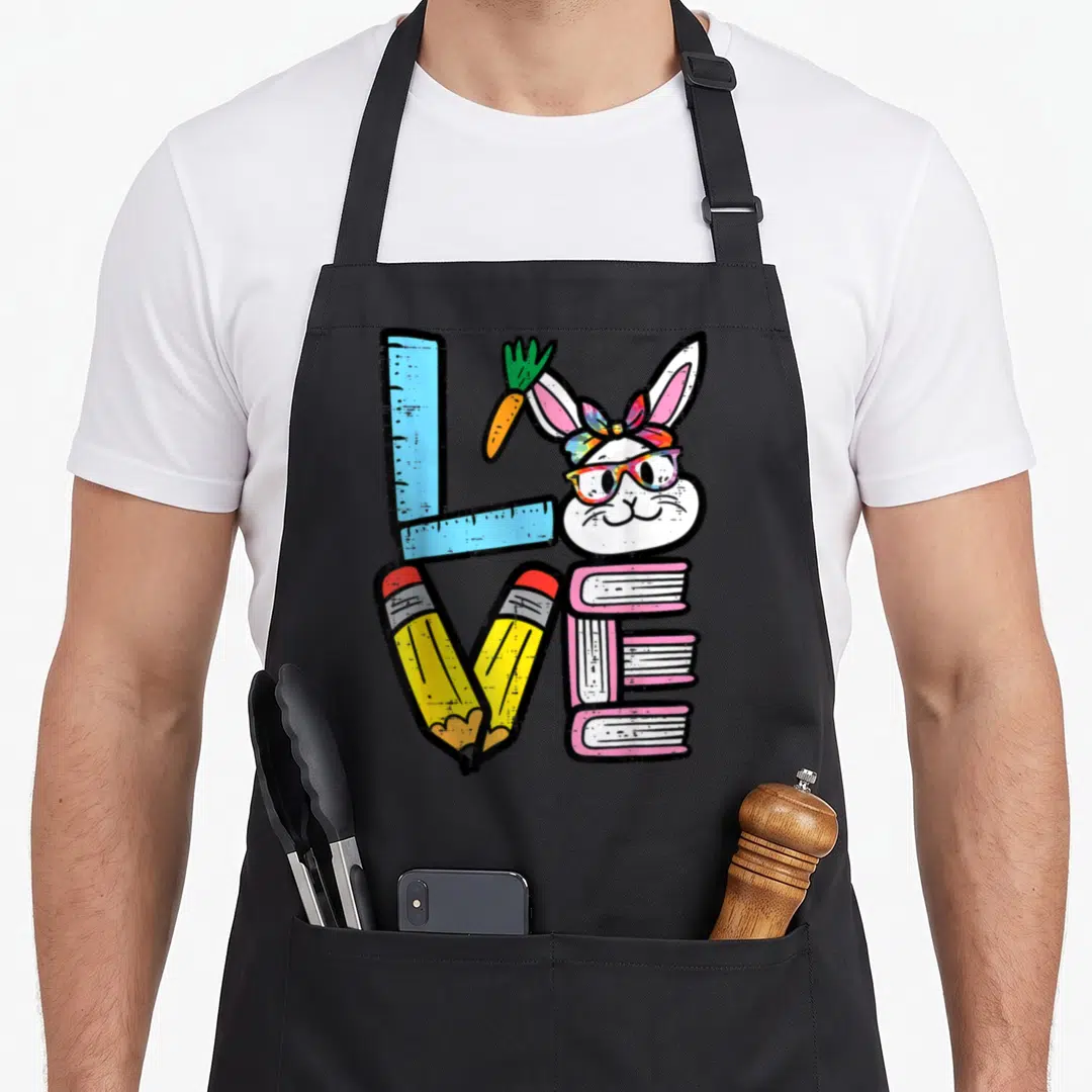 Spring easter bunny aprons - Cartoon bunny head with glasses forms letter O in love spelled by ruler pencils and books — shown on a black cotton canvas apron against a white background
