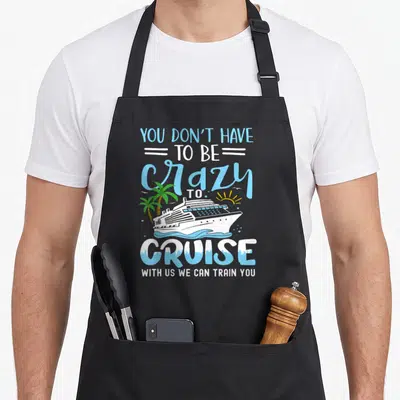 Train us aprons - Cruise ship on blue waves with palm trees and pun text about training to cruise — shown on a black cotton canvas apron against a white background