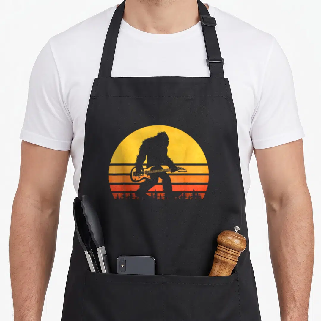 Guitar bigfoot apron - Grey Bigfoot silhouette playing guitar against yellow orange red striped retro sun — shown on a black cotton canvas apron against a white background