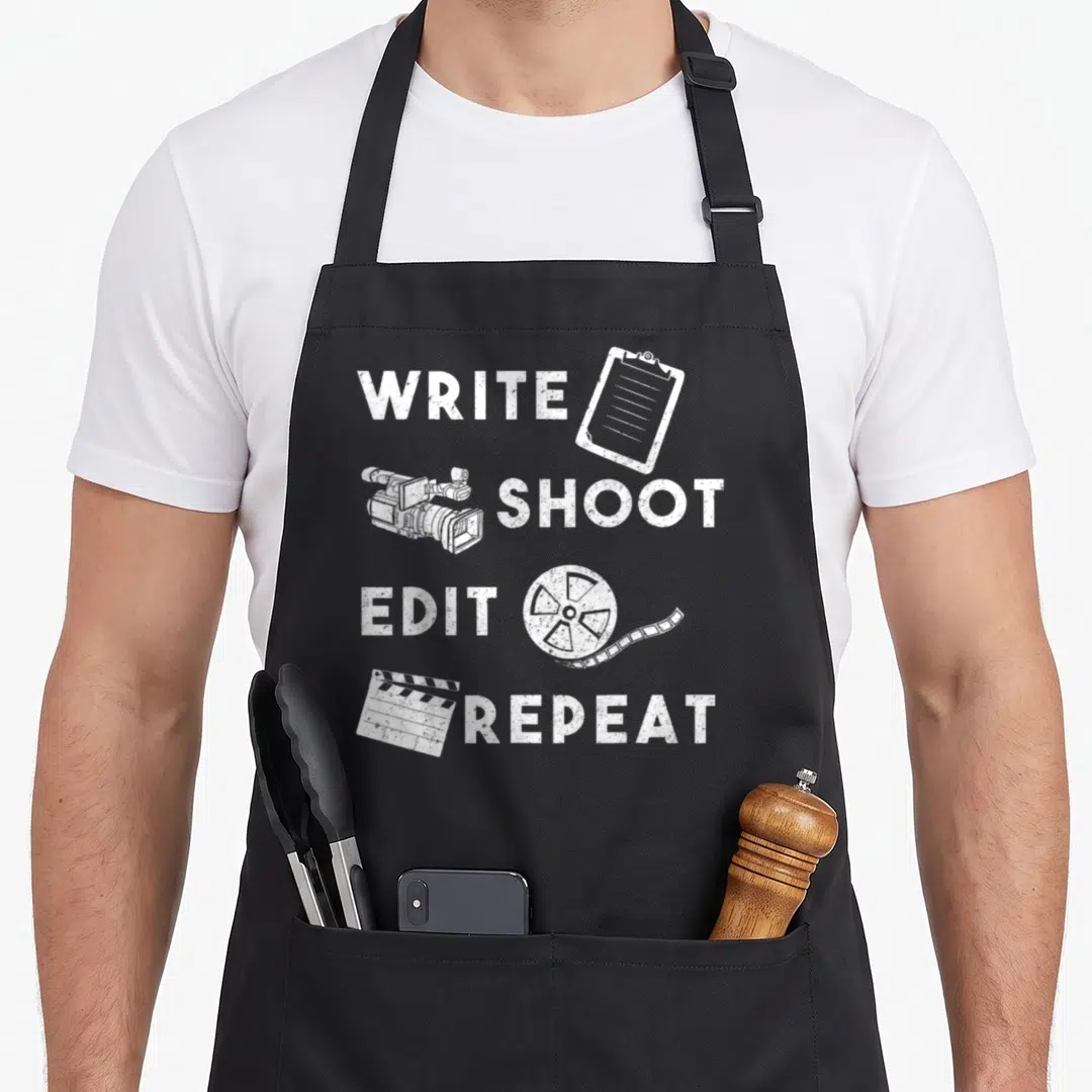 Shoot edit apron - White distressed text Write Shoot Edit Repeat paired with clipboard camera reel and clapperboard — shown on a black cotton canvas apron against a white background