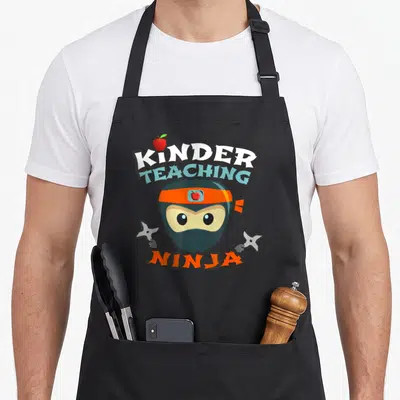 School ninja kindergarten teacher apron - Cartoon ninja head with arched KINDER TEACHING NINJA text, orange headband — shown on a black cotton canvas apron against a white background