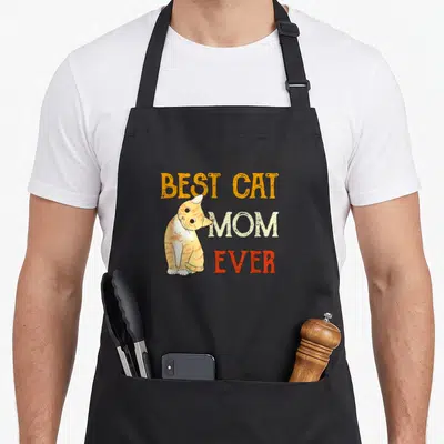 Meow cat in an apron - Cartoon orange tabby cat sitting next to stacked text reading Best Cat Mom Ever in orange white — shown on a black cotton canvas apron against a white background