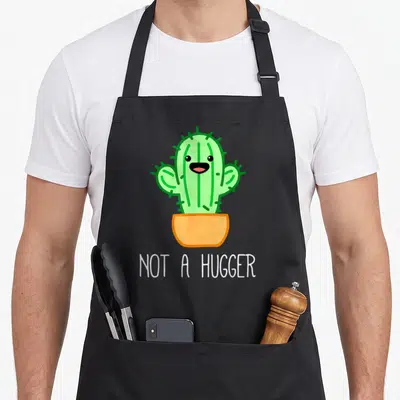 Kawaii cactus apron - Green cartoon cactus in orange pot above white NOT A HUGGER text — shown on a black cotton canvas apron against a white background