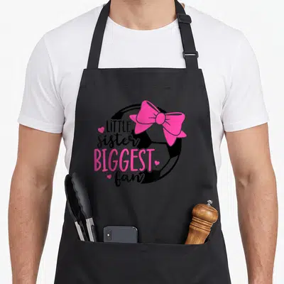 Biggest sister goals apron - LITTLE sister BIGGEST fan text over soccer ball with pink bow and hearts — shown on a black cotton canvas apron against a white background