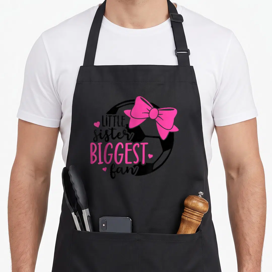 Biggest sister goals apron - LITTLE sister BIGGEST fan text over soccer ball with pink bow and hearts — shown on a black cotton canvas apron against a white background